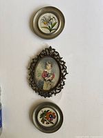Three miniatures arranged on a wall: round daffodil flower in pewter frame, ornate oval boy holding dog painting, and round pansy flower in pewter frame.