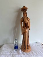 Full view of tall wooden statue of a bearded man wearing a wide-brimmed hat, holding a staff, next to a water bottle for size reference on a white surface against a plain background.