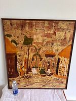 Full view of the framed African batik print showing market scene with traditional houses, people, and palm trees. Includes a water bottle for size reference.