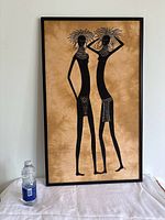 Framed monochrome African batik print showing two young women in tribal jewellery and outfits, set against a warm brown mottled background, with a water bottle for size reference.