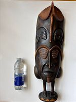 Full front view of tall two-tone ebony African mask with a water bottle for scale, showing detailed faces and feet base.