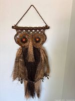 Full view of large owl wall hanging displayed on a white wall, showing entire length and texture, with detail of orange eyes and brown natural fibers.