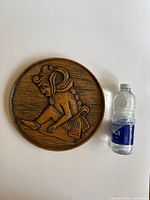 Front view of the round wooden plaque with a detailed carved figure of a Mayan warrior. A water bottle placed beside it for size reference indicates the plaque size is approximately medium-sized.