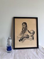 Framed monochrome black and cream print of a Masai mother holding her child, with a water bottle placed beside it for size reference.