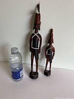 Two African wooden warrior figurines displayed next to a water bottle for size reference. Both figurines show two-tone wood and beaded decorations.