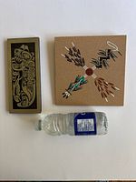 Two indigenous plaques shown side by side with bottle for size reference, Navajo four sacred plants plaque square shaped, Haida etched aluminum rectangular plaque.