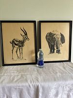 Both framed wildlife prints side by side, one features a gazelle and is signed, the other features an elephant, black ink on creamy paper.