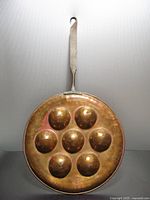 Full view of copper pancake pan showing seven deep concave moulds and long cast iron handle