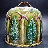 Side view of antique Majolica cheese dome showing raised leaf patterns, rope design arches with pink bows and branch handle on top.