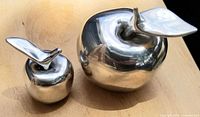 Two polished aluminum apple sculptures on wooden surface, larger and smaller sizes shown