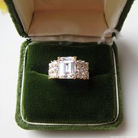 Front view of large emerald cut cubic zirconia centerpiece and surrounding smaller stones in gold plated setting inside green velvet ring case.
