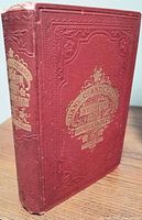 Photo of the front cover showing the red cloth binding, embossed decorative patterns, and gold embossed title text. Some wear visible on edges.