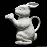 Side view of white bobtail rabbit-shaped ceramic teapot with raised paw as spout and manage on back.