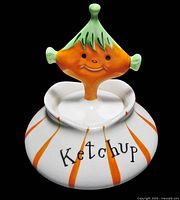 Front view of the vintage white ceramic ketchup jar with orange stripes, 'Ketchup' written on the side, and pixie head lid with green and orange paint.