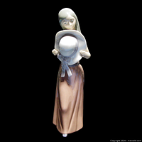 Front view of Lladro Bashful figurine showing girl holding straw hat with ribbon, smooth glazed porcelain finish