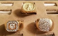 Three gold plated men's rings positioned on a beige textured surface: one ring featuring an embossed classical figure medallion, and two rings with imitation diamond clusters.