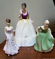 Three Royal Doulton figurines grouped together on table, showing full figures and dress colors