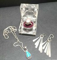 Photo showing the entire group of jewelry: chain with turquoise pendant, large quartz/moonstone ring on a transparent display case, and dangle earrings laid out on a black surface.