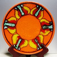 Front view of bright orange round plate with stylized multicolor sunburst interior pattern, displayed on wooden holder.