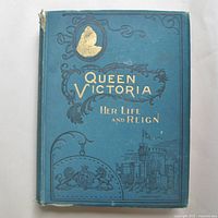 Front cover of the book showing blue background with gold and black embossed title 'Queen Victoria Her Life and Reign', image of Queen Victoria, royal crest and Windsor Castle illustration.