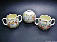 Three item lot showing two double-handled Bunnykins mugs flanking a round Bunnykins bank with stopper, all on dark background.