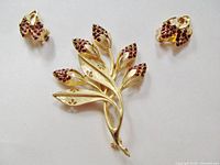Floral brooch and matching clip-on earrings laid flat. Shows front detail of the pieces, yellow gold tone metal with deep red rhinestones clustered on flower buds. Flowing curved stems and textured, shiny finish with leaf cutouts on brooch.