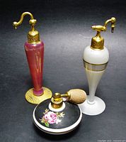 Three perfume atomizers displayed together: one shorter porcelain bottle with floral decoration and two taller glass/brass atomizers.