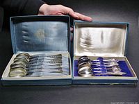 Overview photo showing two boxes open with 6 silver tone teaspoons each inside, visible decorative handle designs and box interiors.