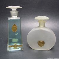 Two 1920s US perfume bottles side by side on grey background, one clear blue rectangular with floral stopper, one frosted oval with dragonfly stopper