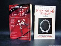Front cover photo showing both paperback books side by side on a black background. Red/black cover on the left is the Official Price Guide to Antique Jewelry, white cover on the right is Collecting Rhinestone Jewelry.