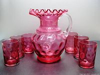 Full set of cranberry glass pitcher and six tumblers arranged in a circle, showcasing color and pattern.