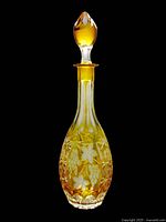 Crystal decanter standing upright, showing golden yellow coloring and grape leaf etching on surface with stopper in place on top.