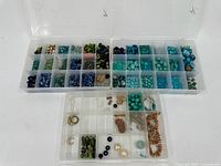 Three plastic organizer trays with many small compartments, each containing various blue and green beads.