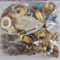 Plastic zipper bag filled with mixed vintage fashion jewelry pieces and craft findings including chains, large beads, feather brooch, and colorful charms.