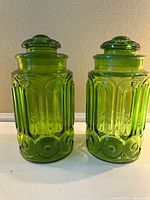 Two green glass jars with lids shown side-by-side, highlighting embossed geometric and circular design patterns on the glass body.