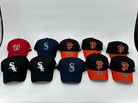 Front view of 10 baseball caps showing distinct team logos and colors, including red Nationals, navy Mariners, black White Sox, and SF Giants caps with orange brims.