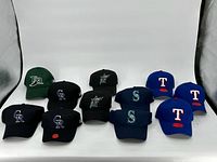 Group of 12 baseball hats arranged in 3 rows on a white backdrop, showing team logos from Major League Baseball.