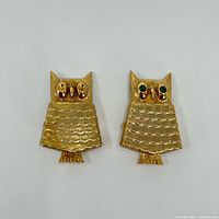 Two gold-tone vintage Avon owl brooches side-by-side on a plain white background, showing front view with one owl having amber gem eyes and the other having green gem eyes.