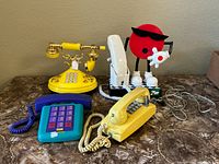 Four plastic landline telephones displayed on a table including yellow rotary-style, blue and purple, yellow push-button phones, and white handset on red 7-Up character base.