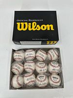 Black Wilson box labeled 'Official Baseballs, One Dozen Baseballs' with 12 baseballs visible inside the box, arranged in 3 rows of 4, each ball wrapped in plastic.
