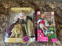 Two boxed Barbie dolls on a brown patterned surface, one Holiday Barbie in gold/purple gown, one Soccer Barbie in sports uniform.