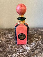 Front view of the pink square glass bottle wrapped in leather, showing the rounded stopper and decorative gold crest emblem on the front.
