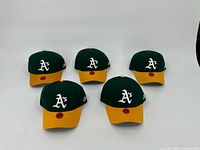 Five Oakland A's baseball hats with green crowns and yellow brims, white embroidered 'A's' logo on front, arranged in a row.