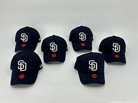 Photo showing all six navy blue San Diego Padres baseball hats with white embroidered SD logos and red S/M size stickers on the brims, arranged in two rows of three on a white surface.