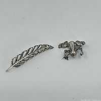 Top view of the sterling silver marcasite frog and leaf brooches lying flat on a white background, showing the detailed texture and design.