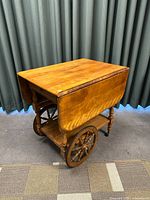 Left angled view of wooden 2-wheeled cart, top leaves extended down, showing two spoked wheels and turned legs.