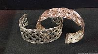 Two braided cuff bracelets displayed side by side on a black background: one silver-tone, the other rose gold plated silver-tone.