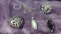 Full lot view showing sterling silver heart locket, sizeable lion ring, green oval pendant on chain, black faceted pendant, and a smaller silver-tone ring.