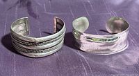 Two sterling silver cuff bracelets displayed side by side on a dark purple surface, one with braided Native American design and the other smooth mirrored finish with some wear.
