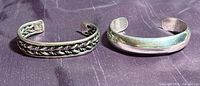 Two sterling silver cuff bracelets placed side by side on purple fabric. One has a braided design, the other is plain with an Indian origin mark.